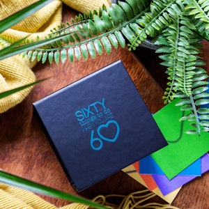 May include: A black box with the text "SIXTY things to do WHEN YOU ARE 60" in blue with a blue heart. The box is on a wooden surface with green ferns and a yellow knitted scarf.
