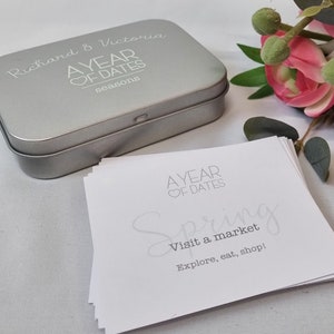 May include: A silver tin box with the text "Richard & Victoria A YEAR OF DATES seasons" on the lid. The box is open and contains a stack of white cards with the text "A YEAR OF DATES Spring Visit a market Explore, eat, shop!" on the top card.