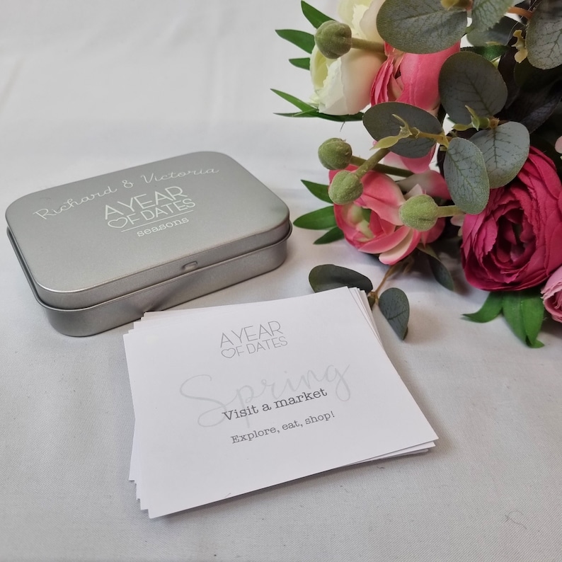 May include: A silver tin box with the text "Richard & Victoria A YEAR OF DATES seasons" on the lid. The box is open and contains a stack of white cards with the text "A YEAR OF DATES Spring Visit a market Explore, eat, shop!" on the top card.