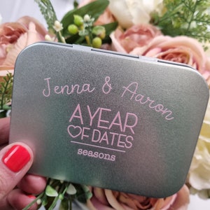 May include: A silver metal tin with the text "Jenna & Aaron A YEAR OF DATES seasons" printed in pink.