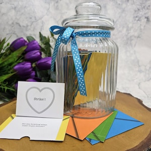 May include: A clear glass jar with a lid, filled with colourful paper strips and decorated with a blue polka dot ribbon. A card with a heart and the word "Strike!!" is in front of the jar, along with origami-style paper shapes.