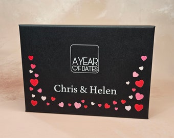 May include: A black box with the text "A YEAR OF DATES" and "Chris & Helen" in white. The box is decorated with red, pink, and white heart shapes. The box is rectangular and appears to be made of cardboard.