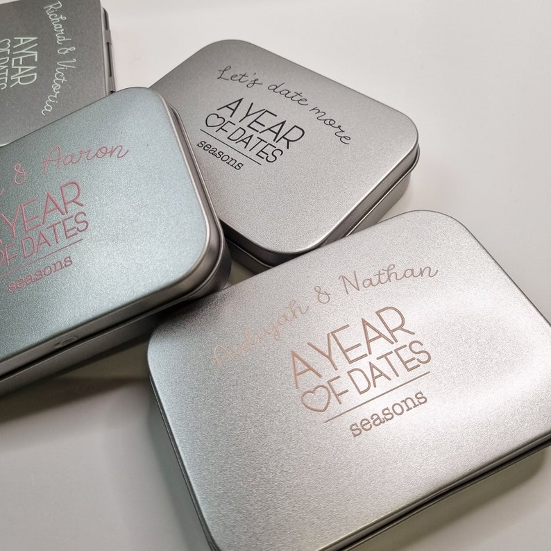 May include: Three silver tins with the text "A Year of Dates" and the words "Seasons" printed on the bottom. The tins are personalized with names and a heart symbol.