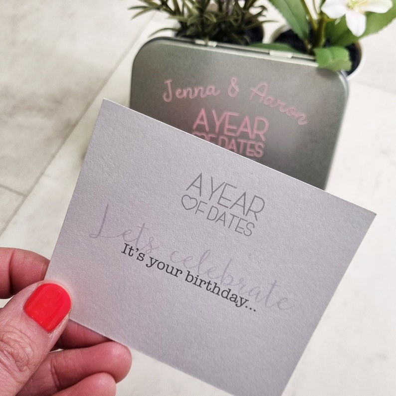 May include: A gray card with the text "Let's celebrate. It's your birthday..." and the logo "A Year of Dates" in pink. The card is in front of a silver tin with the text "Jenna & Aaron A Year of Dates" in pink.