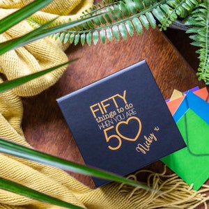 K&ouml;nnte beinhalten: Schwarze Geschenkbox mit goldener Aufschrift "FIFTY things to do when you are 50" mit goldenem Herzen und dem Namen "Nicky x".