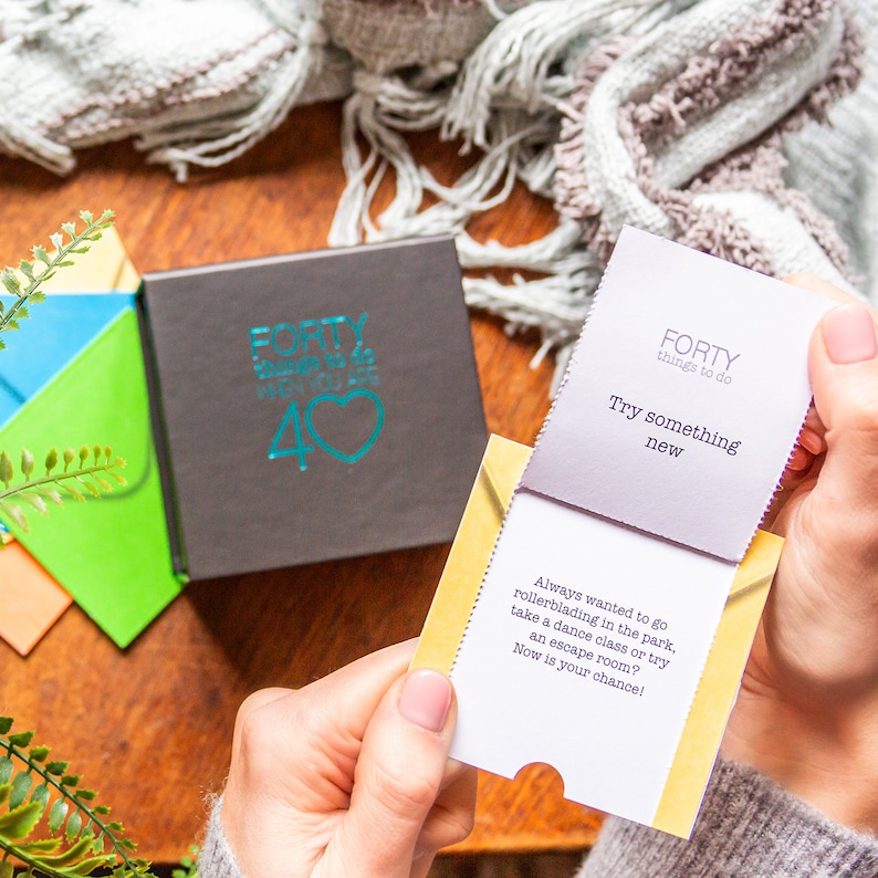 Pu&ograve; includere: Una piccola scatola con un cuore turchese e il testo "FORTY things to do when you are 40" sul davanti. La scatola &egrave; aperta e viene estratta una carta con il testo "FORTY things to do Try something new". La carta &egrave; gialla con un interno bianco e il testo "Always wanted to go rollerblading in the park, take a dance class or try an escape room? Now is your chance!" &egrave; stampato all'interno.