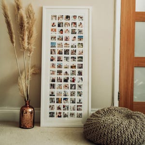 May include: A white picture frame containing a collage of photos. The photos are arranged in a grid pattern and show a variety of people and places. The frame is hanging on a wall with a vase of dried pampas grass in front of it.