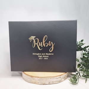 May include: A dark grey graduation certificate holder with gold foil lettering. The text reads "Ruby" with a graduation cap, and "Rivington and Blackrod High School 2017-2022". The holder is displayed on a wooden slice.