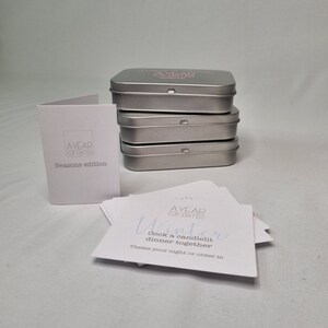 May include: A set of four silver metal tins with the text "A Year of Dates" printed on the top tin. The tins contain a deck of cards with date ideas for each season. The card shown is for winter and reads "Cook a candlelit dinner together. Theme your night or order in."