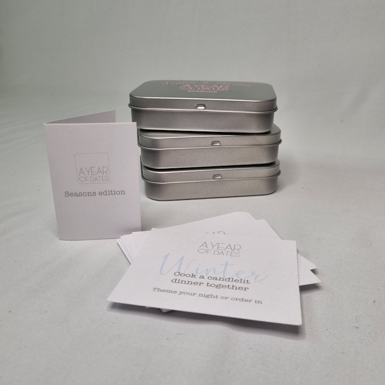 May include: A set of four silver metal tins with the text "A Year of Dates" printed on the top tin. The tins contain a deck of cards with date ideas for each season. The card shown is for winter and reads "Cook a candlelit dinner together. Theme your night or order in."
