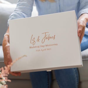 May include: A white memory box with the names "Liz & James" and the text "Wedding Day Memories 25th April 2021" in copper. The box is held by a person wearing a light blue shirt and jeans.