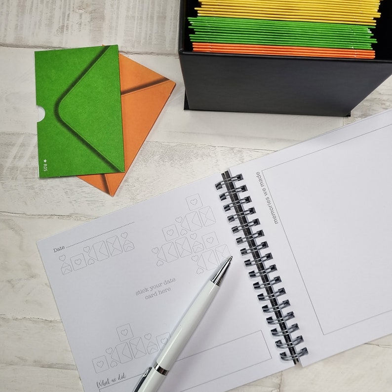 May include: A white spiral bound journal with a gray and white design of envelopes and hearts. The journal has a section for dates and what you did, and a section for memories made. A white pen is lying on the journal.