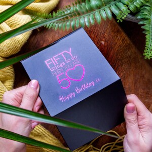 K&ouml;nnte beinhalten: Schwarze Geschenkbox mit einer pinkfarbenen Aufschrift, die "FIFTY things to do when you are 50" mit einem pinkfarbenen Herzen und den Worten "Happy Birthday xx" lautet.