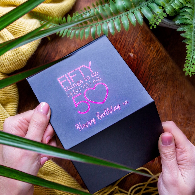 K&ouml;nnte beinhalten: Schwarze Geschenkbox mit einer pinkfarbenen Aufschrift, die "FIFTY things to do when you are 50" mit einem pinkfarbenen Herzen und den Worten "Happy Birthday xx" lautet.