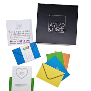 May include: A black box with a silver inscription that reads "A Year of Dates". The box contains 51 small envelopes, each with a different date idea. The envelopes are in various colors, including blue, green, yellow, and orange. The box is a gift for a couple to enjoy a year of dates together.