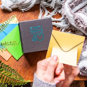Pu&ograve; includere: Una scatola nera con il testo "FORTY Reasons to be 40" in lettere blu turchese e un cuore blu turchese. Una mano tiene una busta gialla con una patta interna bianca.
