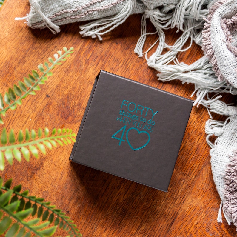 Pu&ograve; includere: Una scatola nera con una scritta blu turchese che recita "FORTY things to do when you are 40" con un cuore blu turchese.