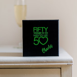 K&ouml;nnte beinhalten: Schwarze Box mit einer gr&uuml;nen Aufschrift, die "FIFTY things to do when you are 50" mit einem gr&uuml;nen Herzsymbol lautet. Der Name "Charles" ist in gr&uuml;ner Kursivschrift unter der Aufschrift geschrieben.