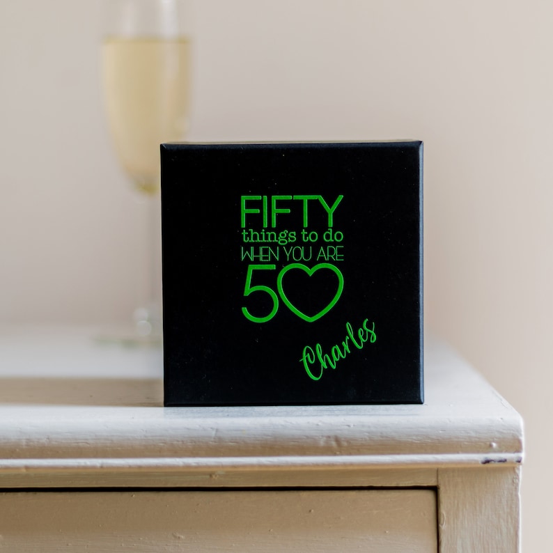 K&ouml;nnte beinhalten: Schwarze Box mit einer gr&uuml;nen Aufschrift, die "FIFTY things to do when you are 50" mit einem gr&uuml;nen Herzsymbol lautet. Der Name "Charles" ist in gr&uuml;ner Kursivschrift unter der Aufschrift geschrieben.