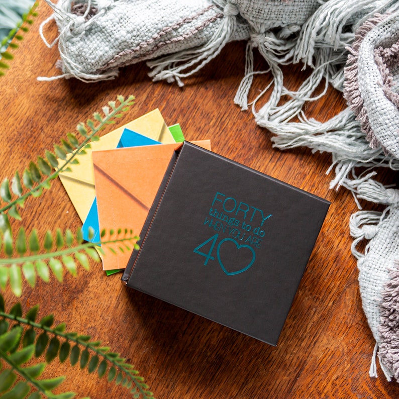 Pu&ograve; includere: Una scatola nera con il testo "FORTY things to do when you are 40" in lettere blu con un cuore blu.