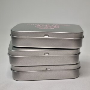 May include: Four silver metal tins stacked on top of each other. The top tin has the text "A YEAR OF DATES" printed in pink on the top.