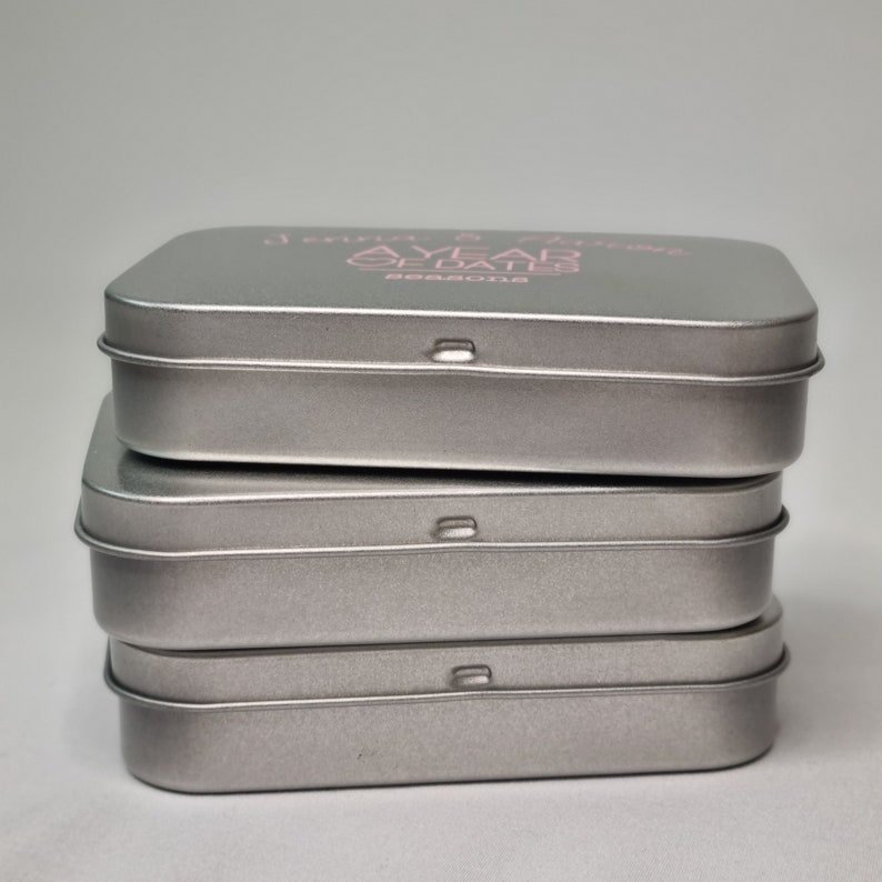 May include: Four silver metal tins stacked on top of each other. The top tin has the text "A YEAR OF DATES" printed in pink on the top.