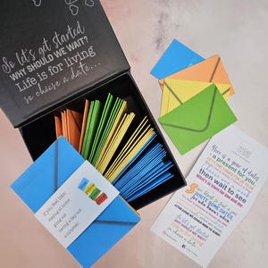 May include: A black box with a lid open, revealing a stack of colorful envelopes. The box is labeled "So let's get started WHY SHOULD WE WAIT? Life is for living so choose a date..."  The envelopes are blue, yellow, green, and orange. A card with the text "Here is a year of dates - a present for you. FIFTY ONE THINGS FOR US TO DO Choose one a week then wait to see What's in store for you and me." is visible in the background.