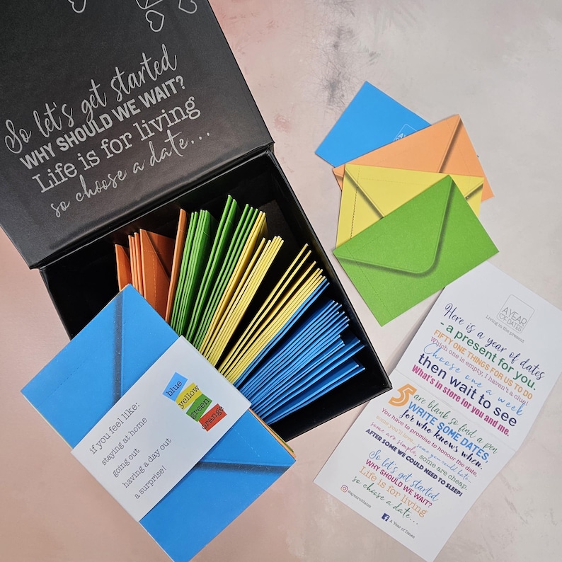 May include: A black box with a lid open, revealing a stack of colorful envelopes. The box is labeled "So let's get started WHY SHOULD WE WAIT? Life is for living so choose a date..."  The envelopes are blue, yellow, green, and orange. A card with the text "Here is a year of dates - a present for you. FIFTY ONE THINGS FOR US TO DO Choose one a week then wait to see What's in store for you and me." is visible in the background.