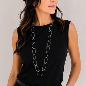 Può includere: Una lunga collana color argento caratterizzata da una serie di maglie metalliche ovali interconnesse. La catena è composta da sottili anelli circolari e ovali lisci di varie dimensioni che pendono verticalmente su un top nero senza maniche.