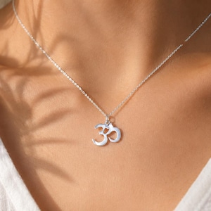 Könnte beinhalten: Eine silberfarbene Halskette mit einer feinen Kette und einem kleinen, polierten Anhänger in Form des Om-Symbols. Der Anhänger ist mittig an der Kette platziert, die am Schlüsselbein aufliegt. Das Metall hat ein helles, reflektierendes Finish.