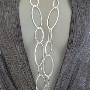 Könnte beinhalten: Eine silberfarbene Halskette mit einer Reihe von miteinander verbundenen ovalen Gliedern. Die Kette wird vor einem dunklen, strukturierten Hintergrund präsentiert, der das polierte Metall hervorhebt. Das Design ist modern und elegant.