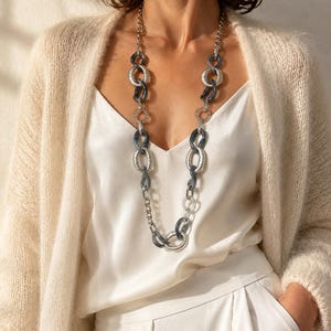 Puede incluir: Un collar largo que presenta eslabones ovalados alternos en metal plateado y resina gris oscuro translúcida. El diseño de la cadena incorpora anillos metálicos texturizados y bucles de resina lisa y brillante, creando un aspecto superpuesto.