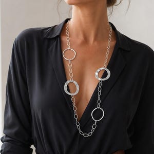 Può includere: Una lunga collana color argento caratterizzata da una serie di maglie ovali interconnesse e tre anelli circolari più grandi in metallo spazzolato distanziati lungo la catena. La collana pende verticalmente su una camicia nera con colletto.