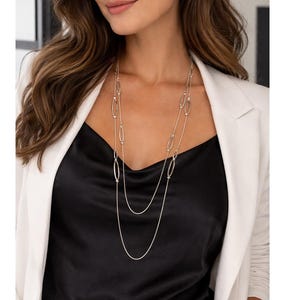 May include: Silver necklace featuring oval links and a fine chain, layered over a black satin top. The model wears a white blazer and small hoop earrings. The necklace is a stylish accessory.