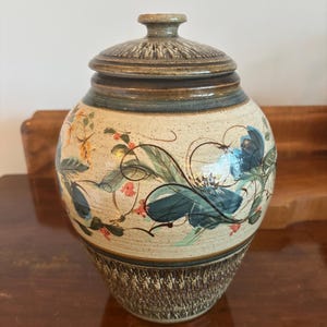 May include: A ceramic jar with a lid. The beige body is decorated with blue, green, and orange floral patterns. The jar has a textured, dark brown base and a matching lid. The design is rustic and artistic, suitable for home decor.