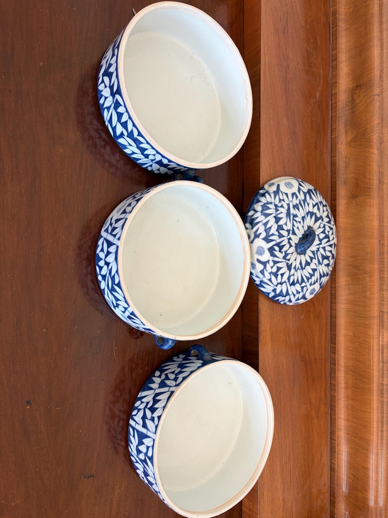 Puede incluir: Tres cuencos redondos de cer&aacute;mica con interiores blancos y patrones bot&aacute;nicos azules en los lados exteriores. Una tapa circular a juego con un dise&ntilde;o de hojas azul y blanco se coloca a la derecha de los cuencos. Cada cuenco tiene peque&ntilde;as asas laterales.