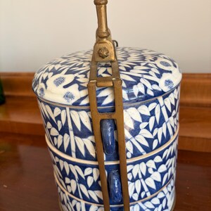 Puede incluir: Un recipiente cil&iacute;ndrico de porcelana de tres niveles con un patr&oacute;n azul y blanco de bamb&uacute; y flores. Las secciones apilables est&aacute;n aseguradas por un marco vertical de lat&oacute;n con un asa central que se extiende desde la tapa.