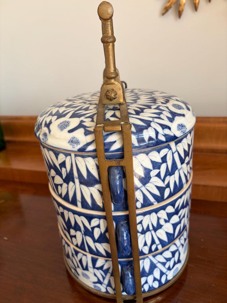 Puede incluir: Un recipiente cil&iacute;ndrico de porcelana de tres niveles con un patr&oacute;n azul y blanco de bamb&uacute; y flores. Las secciones apilables est&aacute;n aseguradas por un marco vertical de lat&oacute;n con un asa central que se extiende desde la tapa.