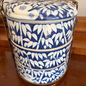 Puede incluir: Un portacomidas de cer&aacute;mica de tres niveles con un patr&oacute;n azul y blanco de bamb&uacute; y flores. Los recipientes cil&iacute;ndricos est&aacute;n apilados verticalmente y asegurados por un marco de lat&oacute;n con un asa que se arquea sobre la tapa superior.