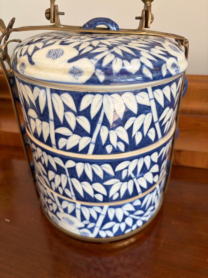 Puede incluir: Un portacomidas de cer&aacute;mica de tres niveles con un patr&oacute;n azul y blanco de bamb&uacute; y flores. Los recipientes cil&iacute;ndricos est&aacute;n apilados verticalmente y asegurados por un marco de lat&oacute;n con un asa que se arquea sobre la tapa superior.