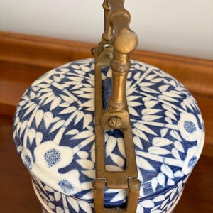 Puede incluir: Un recipiente cil&iacute;ndrico de cer&aacute;mica con un patr&oacute;n floral y de bamb&uacute; en azul y blanco. El objeto presenta un dise&ntilde;o escalonado sujeto por un marco vertical de lat&oacute;n con un asa texturizada que se arquea sobre la tapa superior.