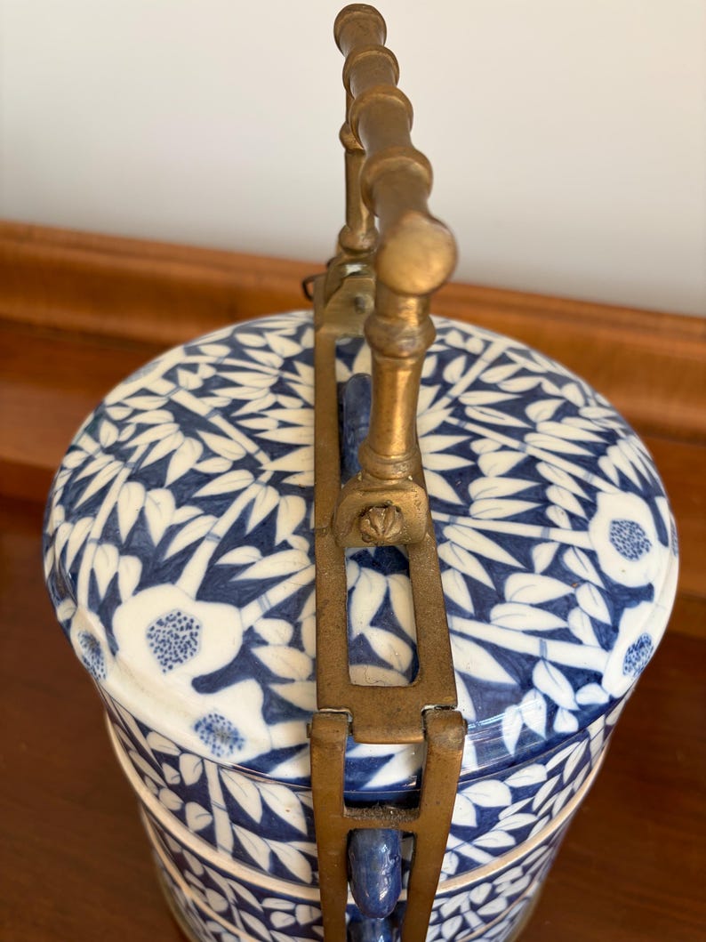 Puede incluir: Un recipiente cil&iacute;ndrico de cer&aacute;mica con un patr&oacute;n floral y de bamb&uacute; en azul y blanco. El objeto presenta un dise&ntilde;o escalonado sujeto por un marco vertical de lat&oacute;n con un asa texturizada que se arquea sobre la tapa superior.
