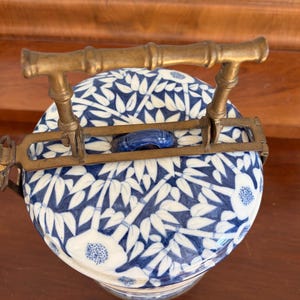 Puede incluir: Un recipiente redondo de cer&aacute;mica con un patr&oacute;n de bamb&uacute; y flores en azul y blanco, que presenta un asa de metal de lat&oacute;n con forma de tallos de bamb&uacute;. La tapa tiene un peque&ntilde;o pomo de cer&aacute;mica azul y los herrajes incluyen un mecanismo.
