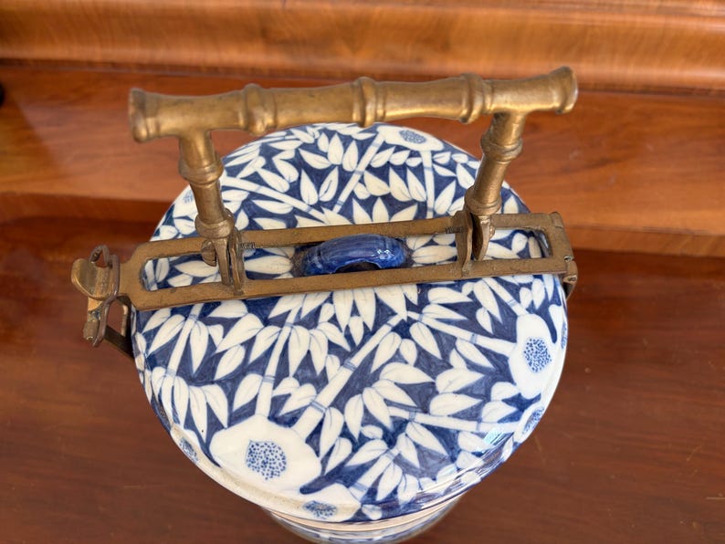 Puede incluir: Un recipiente redondo de cer&aacute;mica con un patr&oacute;n de bamb&uacute; y flores en azul y blanco, que presenta un asa de metal de lat&oacute;n con forma de tallos de bamb&uacute;. La tapa tiene un peque&ntilde;o pomo de cer&aacute;mica azul y los herrajes incluyen un mecanismo.