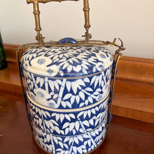 Puede incluir: Un portacomidas cil&iacute;ndrico de tres niveles hecho de porcelana blanca con un patr&oacute;n de hojas de bamb&uacute; azul. El recipiente cuenta con un marco de metal de lat&oacute;n y un asa con forma de bamb&uacute;, con un mecanismo de cierre para los niveles.