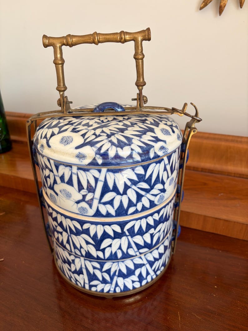 Puede incluir: Un portacomidas cil&iacute;ndrico de tres niveles hecho de porcelana blanca con un patr&oacute;n de hojas de bamb&uacute; azul. El recipiente cuenta con un marco de metal de lat&oacute;n y un asa con forma de bamb&uacute;, con un mecanismo de cierre para los niveles.