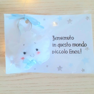 Può includere: Un portachiavi di peluche bianco a forma di nuvola con un fiocco blu e un viso sorridente. Il portachiavi è attaccato a una carta bianca con il testo "Benvenuto in questo mondo piccolo Enea!" in nero.