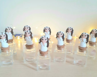 May include: A collection of small glass jars with cork lids, each topped with a white figurine of a girl wearing a white dress and a white flower crown. The figurines are all in a praying position.