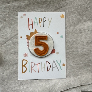 Puede incluir: Una tarjeta de cumpleaños con las palabras "HAPPY BIRTHDAY" en letras coloridas. La tarjeta presenta el número "5" de fieltro naranja, adjunto a un círculo blanco, con una ilustración de corona. Pequeñas ilustraciones de flores están dispersas en la tarjeta.