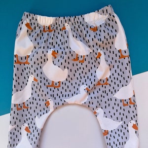 A pair of child harem pants on a blue and white background. Pant fabric print has a grey background with black dashes and a white duck with orange beak and feet.